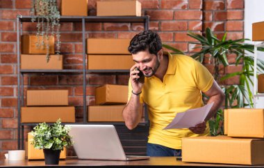 Hintli Asyalı Adam Dizüstü bilgisayarlı ve akıllı telefonlu online siparişleri yönetiyor, Depo 'dan gönderilmeye hazır.