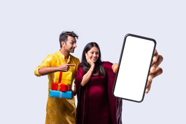 Etnik kökenli Hintli genç çift ellerinde boş ekran ve alışveriş torbalarıyla büyük bir akıllı telefon tutuyorlar.