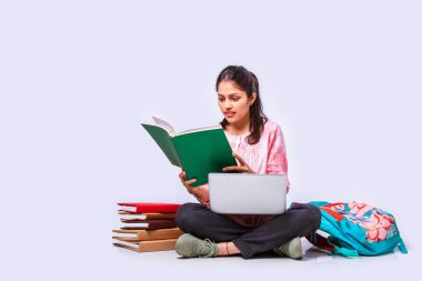 Hintli Asyalı üniversite öğrencisi dizüstü bilgisayarla beyaz arka planda otururken okuyor.