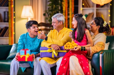 Aile Diwali gecesini kutluyor, ladaları paylaşıyor ve hediyelerle dolu bir kanepede neşe saçıyor.