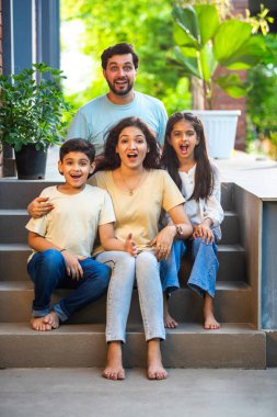 Dört çocuklu gülümseyen Hintli ailenin portresi, genç ebeveynler ve iki çocuk, neşeli bir şekilde evlerinin önünde oturuyorlar, birlikte sıcak ve mutlu bir anı paylaşıyorlar, aşkı kutluyorlar, aile bağlarını.