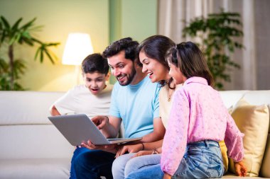 Dört kişilik Hintli Asyalı genç aile bir koltukta oturmuş, eğlence veya öğrenim için tablet ya da dizüstü bilgisayar kullanarak teknolojiyle uğraşıyorlar.