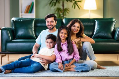 Dört kişilik gülümseyen Hintli Asyalı aile, aileleri sevgiyle kucağında kız ve oğullarıyla ya da çocuklar şık, modern bir evin içinde, bir portre için poz veriyorlar.