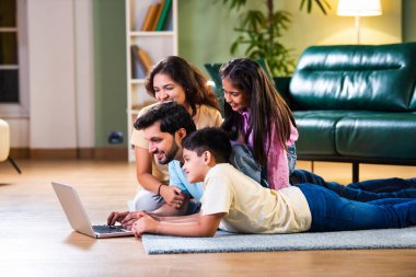 Dört kişilik Hintli Asyalı genç aile bir koltukta oturmuş, eğlence veya öğrenim için tablet ya da dizüstü bilgisayar kullanarak teknolojiyle uğraşıyorlar.