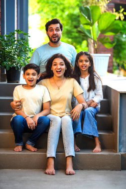 Dört çocuklu gülümseyen Hintli ailenin portresi, genç ebeveynler ve iki çocuk, neşeli bir şekilde evlerinin önünde oturuyorlar, birlikte sıcak ve mutlu bir anı paylaşıyorlar, aşkı kutluyorlar, aile bağlarını.
