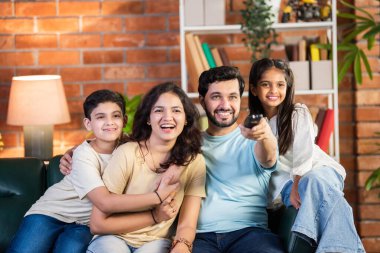 Dört kişilik neşeli Hintli aile oturma odasındaki kanepede oturup uzaktan kumandayla televizyon izleyip kaliteli bir aile ve eğlence ortamının tadını çıkarıyorlar.