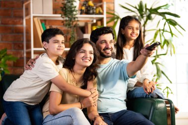 Dört kişilik neşeli Hintli aile oturma odasındaki kanepede oturup uzaktan kumandayla televizyon izleyip kaliteli bir aile ve eğlence ortamının tadını çıkarıyorlar.