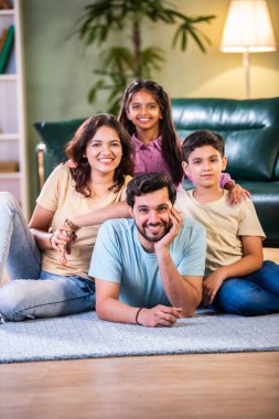 Neşeli Hintli Asyalı aile evde eğleniyor. Kocası çenesinde ellerle yerde yatıyor. Eşi yanında oturuyor ve çocuklar sırtüstü oturuyor.