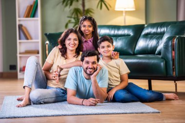 Neşeli Hintli Asyalı aile evde eğleniyor. Kocası çenesinde ellerle yerde yatıyor. Eşi yanında oturuyor ve çocuklar sırtüstü oturuyor.