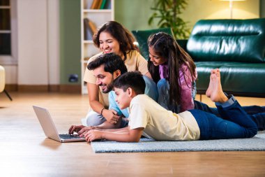 Dört kişilik Hintli Asyalı genç aile bir koltukta oturmuş, eğlence veya öğrenim için tablet ya da dizüstü bilgisayar kullanarak teknolojiyle uğraşıyorlar.