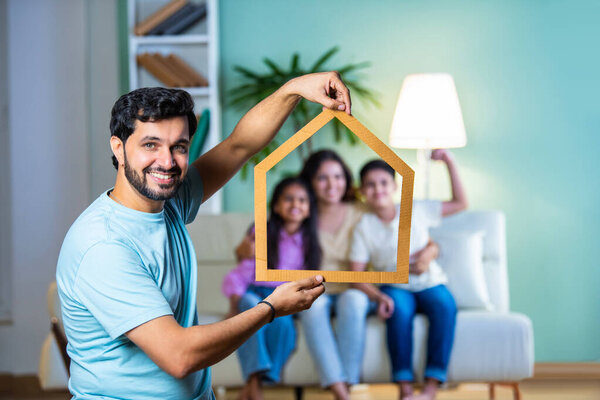 Индийский молодой отец-азиат держит на заднем плане картонную фигуру в форме дома с семьей, сидящей на диване, символизируя мечты о недвижимости и домовладение