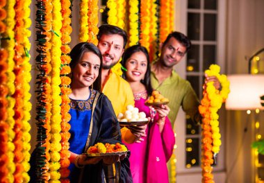 Neşeli Hintli Asyalı iki arkadaş geleneksel kıyafetleriyle Diwali 'yi kutluyorlar evde, kadife çiçekleri ve şekerlerle dolu bir tabağın içinden neşeyle gözetliyorlar.