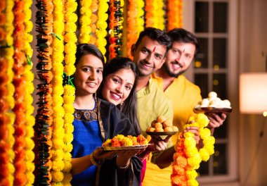 Neşeli Hintli Asyalı iki arkadaş geleneksel kıyafetleriyle Diwali 'yi kutluyorlar evde, kadife çiçekleri ve şekerlerle dolu bir tabağın içinden neşeyle gözetliyorlar.