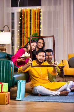 Dört ya da genç çiftten oluşan neşeli Hintli aile Diwali gecesinin tadını çıkarıyor, rahat bir koltukta oturuyor, boş bir çerçeveden neşeyle bakıyor, süslü bir evde şenlikli anları yakalıyor.