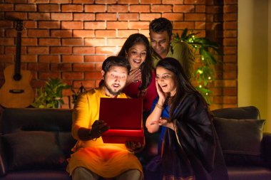 Hintli aile veya genç çiftler Diwali 'yi evde kutluyorlar, koltukta oturuyorlar, parlayan sürpriz bir hediye kutusu açıyorlar, Hindu bayramı boyunca birlikte neşe ve heyecan paylaşıyorlar.