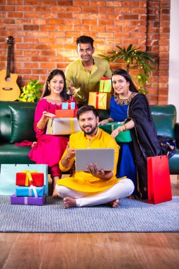 Geleneksel giysiler içinde neşeli Hintli çiftler Diwali 'yi evde kutluyorlar, kanepeye oturmuş tatlı, hediye ve çiçeklerle gülümsüyorlar, online alışveriş veya video çağrısı için dizüstü bilgisayar kullanıyorlar.