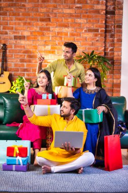 Geleneksel giysiler içinde neşeli Hintli çiftler Diwali 'yi evde kutluyorlar, kanepeye oturmuş tatlı, hediye ve çiçeklerle gülümsüyorlar, online alışveriş veya video çağrısı için dizüstü bilgisayar kullanıyorlar.