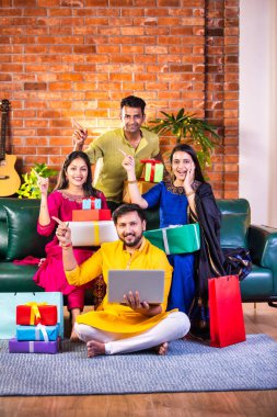 Geleneksel giysiler içinde neşeli Hintli çiftler Diwali 'yi evde kutluyorlar, kanepeye oturmuş tatlı, hediye ve çiçeklerle gülümsüyorlar, online alışveriş veya video çağrısı için dizüstü bilgisayar kullanıyorlar.