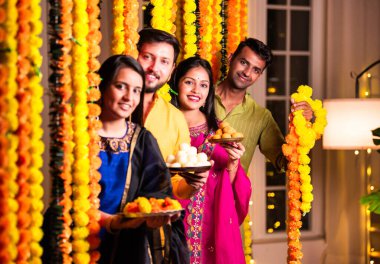 Neşeli Hintli Asyalı iki arkadaş geleneksel kıyafetleriyle Diwali 'yi kutluyorlar evde, kadife çiçekleri ve şekerlerle dolu bir tabağın içinden neşeyle gözetliyorlar.