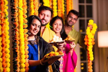 Neşeli Hintli Asyalı iki arkadaş geleneksel kıyafetleriyle Diwali 'yi kutluyorlar evde, kadife çiçekleri ve şekerlerle dolu bir tabağın içinden neşeyle gözetliyorlar.