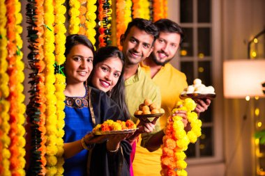 Neşeli Hintli Asyalı iki arkadaş geleneksel kıyafetleriyle Diwali 'yi kutluyorlar evde, kadife çiçekleri ve şekerlerle dolu bir tabağın içinden neşeyle gözetliyorlar.