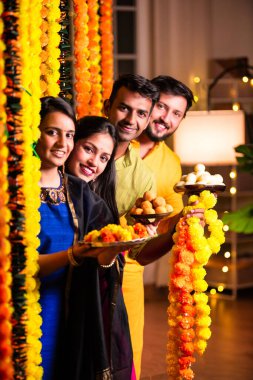 Neşeli Hintli Asyalı iki arkadaş geleneksel kıyafetleriyle Diwali 'yi kutluyorlar evde, kadife çiçekleri ve şekerlerle dolu bir tabağın içinden neşeyle gözetliyorlar.