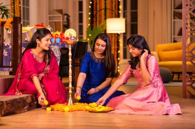 Joyful Indian Asian young girls or sisters in traditional wear creating vibrant flower rangoli around a traditional Samai or diya, lighting it to celebrate Diwali in luxurious home interiors clipart