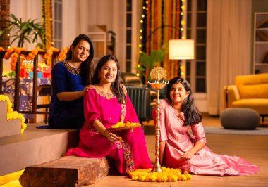 Joyful Indian Asian young girls or sisters in traditional wear creating vibrant flower rangoli around a traditional Samai or diya, lighting it to celebrate Diwali in luxurious home interiors clipart