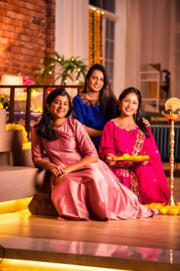 Joyful Indian Asian young girls or sisters in traditional wear creating vibrant flower rangoli around a traditional Samai or diya, lighting it to celebrate Diwali in luxurious home interiors clipart