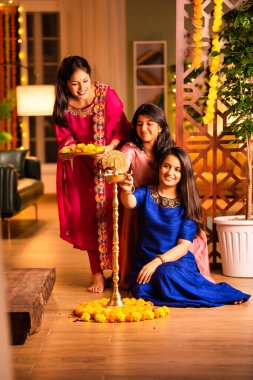 Joyful Indian Asian young girls or sisters in traditional wear creating vibrant flower rangoli around a traditional Samai or diya, lighting it to celebrate Diwali in luxurious home interiors clipart