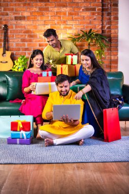 Geleneksel giysiler içinde neşeli Hintli çiftler Diwali 'yi evde kutluyorlar, kanepeye oturmuş tatlı, hediye ve çiçeklerle gülümsüyorlar, online alışveriş veya video çağrısı için dizüstü bilgisayar kullanıyorlar.