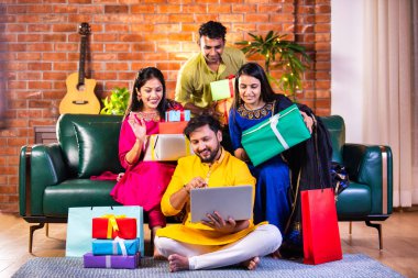 Geleneksel giysiler içinde neşeli Hintli çiftler Diwali 'yi evde kutluyorlar, kanepeye oturmuş tatlı, hediye ve çiçeklerle gülümsüyorlar, online alışveriş veya video çağrısı için dizüstü bilgisayar kullanıyorlar.