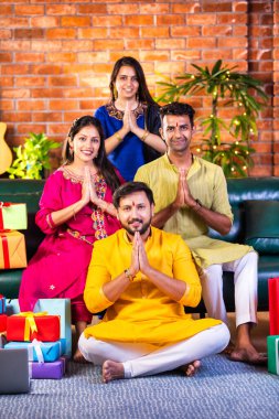 Joyful Indian Asian couples in traditional attire celebrating Diwali at home, sitting on a sofa, greeting guests with Namaste or Namaskar, enjoying the festive occasion together with love and respect clipart