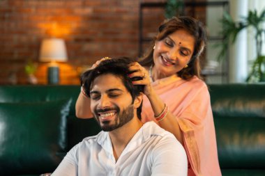 60 'lı yaşlardaki yaşlı Hintli anne, genç ve yakışıklı oğluna keyifsiz ışıklandırmalı, rahat, modern bir oturma odasında yağ masajı yaparak rahatlatıcı ve besleyici bir atmosfer yaratıyor.