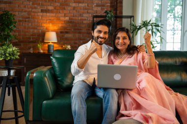 60 'lı yaşlarda yaşlı Hintli bir anne ve yakışıklı oğlu online alışveriş veya video çağrısı için dizüstü bilgisayar kullanıyorlar. Sıcak, kasvetli ışıklandırması olan modern bir oturma odasında oturuyorlar.