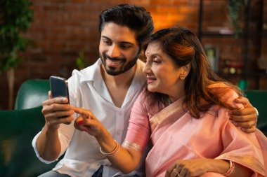 60 'lı yaşlarda Hintli bir anne, genç ve yakışıklı oğluyla birlikte bir koltukta otururken ya da modern bir oturma odasında karamsar ışıklandırması olan bir masada otururken akıllı telefonuyla fotoğraf ya da video izlerdi.