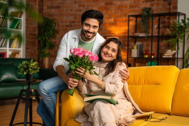 Neşeli yaşlı Hintli anne, Anneler Günü 'nde küçük oğluyla birlikte sıcak, karamsar bir kanepede otururken neşeyle hediyelerini ve güzel bir çiçek buketini değiş tokuş ediyor.