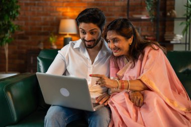 60 'lı yaşlarda yaşlı Hintli bir anne ve yakışıklı oğlu online alışveriş veya video çağrısı için dizüstü bilgisayar kullanıyorlar. Sıcak, kasvetli ışıklandırması olan modern bir oturma odasında oturuyorlar.