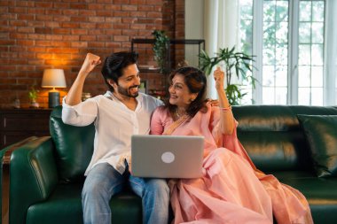 60 'lı yaşlarda yaşlı Hintli bir anne ve yakışıklı oğlu online alışveriş veya video çağrısı için dizüstü bilgisayar kullanıyorlar. Sıcak, kasvetli ışıklandırması olan modern bir oturma odasında oturuyorlar.