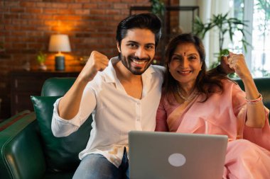 60 'lı yaşlarda yaşlı Hintli bir anne ve yakışıklı oğlu online alışveriş veya video çağrısı için dizüstü bilgisayar kullanıyorlar. Sıcak, kasvetli ışıklandırması olan modern bir oturma odasında oturuyorlar.