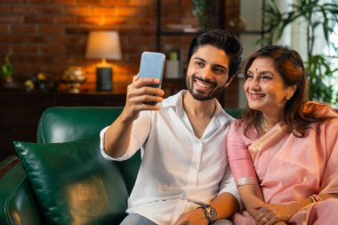 60 'lı yaşlarda Hintli bir anne, genç ve yakışıklı oğluyla birlikte bir koltukta otururken ya da modern bir oturma odasında karamsar ışıklandırması olan bir masada otururken akıllı telefonuyla fotoğraf ya da video izlerdi.