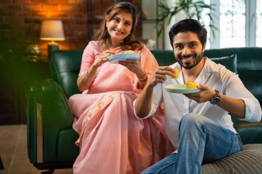 Altmışlarında Hintli bir anne ve genç ve yakışıklı oğlu sıcak, karamsar, modern oturma odasındaki rahat bir kanepede sohbet ederken geleneksel bir dhokla aperatifinin ya da hamur işinin tadını çıkarıyorlar.