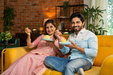 Altmışlarında Hintli bir anne ve genç ve yakışıklı oğlu sıcak, karamsar, modern oturma odasındaki rahat bir kanepede sohbet ederken geleneksel bir dhokla aperatifinin ya da hamur işinin tadını çıkarıyorlar.