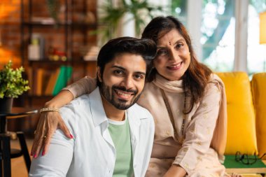 Sari içinde yaşlı bir Hintli annenin portresi yakışıklı oğlunu kucaklarken sıcak, karamsar aydınlatmalarla çevrili rahat bir kanepede otururken sevgi dolu bağlarını güzel bir şekilde ön plana çıkarıyor.