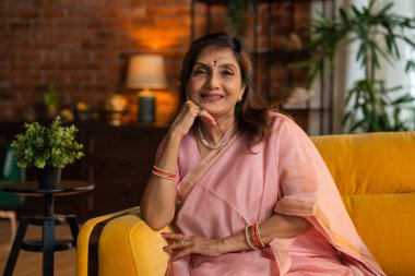 Göz kamaştırıcı bir sari içinde yaşlı, güzel bir Hintli kadının portresi, bir kanepede rahat bir şekilde otururken kendine güven ve sükunet yayıyor, karamsar bir ışıkta kraliyet zarafetinin havasını yakalıyor.