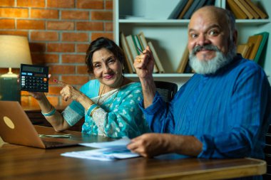 Asyalı Hintli yaşlı çift bir dizüstü bilgisayar, kâğıt, hesap makinesi ve para vantilatörü kullanarak evdeki doğru muhasebeye odaklanarak masalarında ev finansmanını özenle yönetiyor.