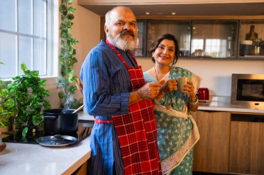 Charming Indian Asian senior couple enjoying a warm cup of tea with biscuits while waiting for their meal to be cooked in a beautifully modern and well-equipped kitchen clipart