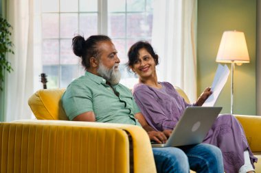 Hintli orta yaşlı çift sarı koltukta oturuyor, sakallı koca, bilgisayarını muhasebe için kullanıyor ve belgelere bakıyor, canlı, rahat bir evde.