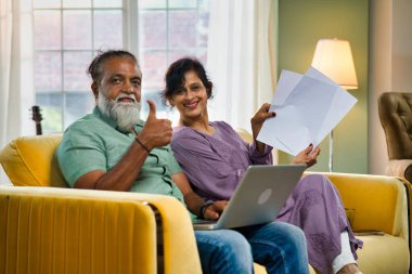 Hintli orta yaşlı çift sarı koltukta oturuyor, sakallı koca, bilgisayarını muhasebe için kullanıyor ve belgelere bakıyor, canlı, rahat bir evde.