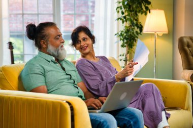 Hintli orta yaşlı çift sarı koltukta oturuyor, sakallı koca, bilgisayarını muhasebe için kullanıyor ve belgelere bakıyor, canlı, rahat bir evde.